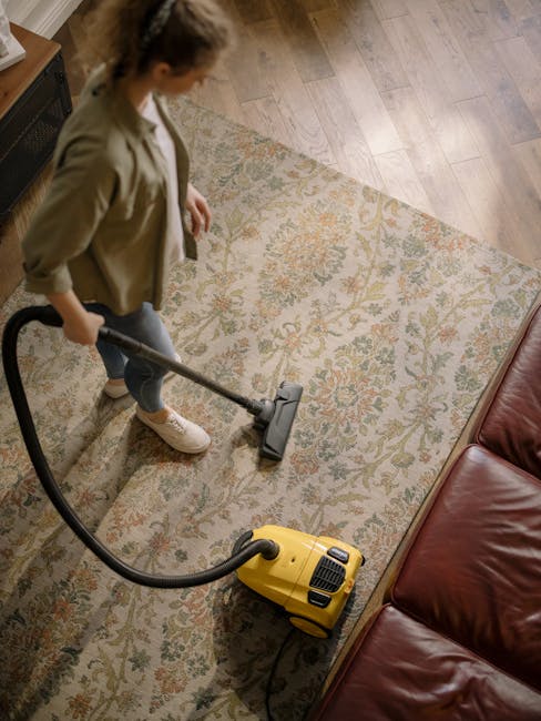 A person kneeling on a patterned carpet in a room with warm lighting, loading a white cloth into a yellow and black vacuum cleaner with a flexible hose, indicating equipment used for surface cleaning 