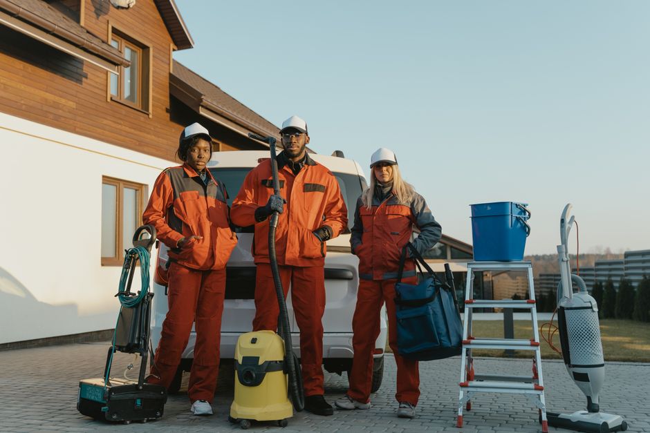 Three professional cleaning specialists dressed in orange and gray uniforms with white safety helmets standing outdoors in front of a white delivery van. They are equipped with various cleaning tools,