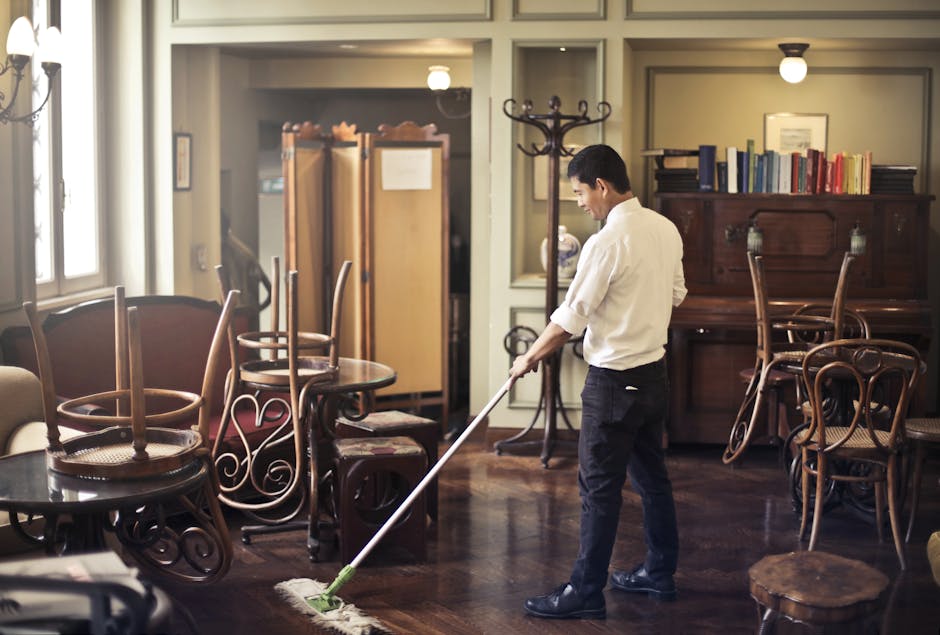 A professional cleaner from crystalpalacecarpetcleaners.co.uk indoors, holding a spray bottle of cleaning solution in her right hand and a cleaning brush with a blue sponge head in her left hand. She 